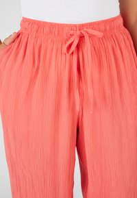 Pantaloni crepe color corallo con cintura elastica e cordoncino. Caratterizzati da tasche laterali e un tessuto leggero e testurizzato.