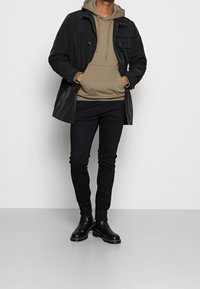 Chubasquero negro impermeable con múltiples bolsillos frontales sobre una sudadera beige. Pantalones negros de corte slim y botas negras hasta el tobillo completan el conjunto.