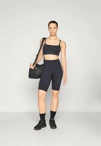 Fekete sportmelltartó és magas derekú kerékpáros nadrág, mindkettő varrás nélküli kialakításban. A modell fekete tornazsákot tart, fekete sportcipőt és zoknit visel.