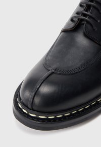 Schwarzer Lederschuh mit abgerundeter Zehenpartie, sichtbaren Nähdetails, einer Gummisohle und schwarz-weißen Nähten entlang der Sohlenkante.