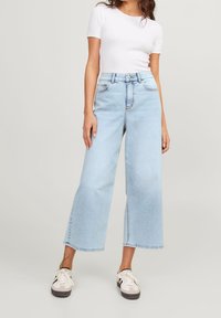 Kvinna som bär ljusblå vida caprijeans, vit tight t-shirt med kort ärm och vita sneakers med svarta detaljer, stående.