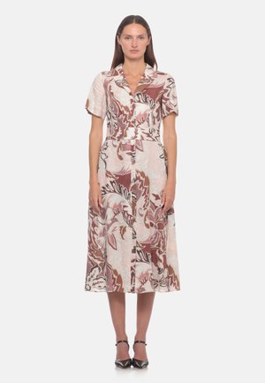 Donna in piedi che indossa un vestito midi con maniche corte e un motivo floreale astratto marrone, rosa e bianco, abbinato a tacchi neri con cinturino alla caviglia.