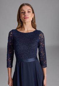 Abito da sera blu navy con corpetto in pizzo con maniche a tre quarti, fascia in satin liscia in vita e gonna fluente.