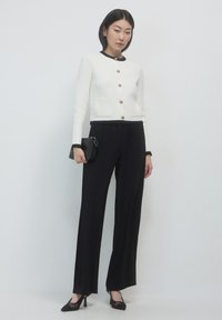 Weißer Cardigan mit goldenen Knöpfen und schwarzem Besatz, kombiniert mit schwarzen, weit geschnittenen Hosen. Das Model hält eine kleine schwarze Handtasche und trägt schwarze High Heels.