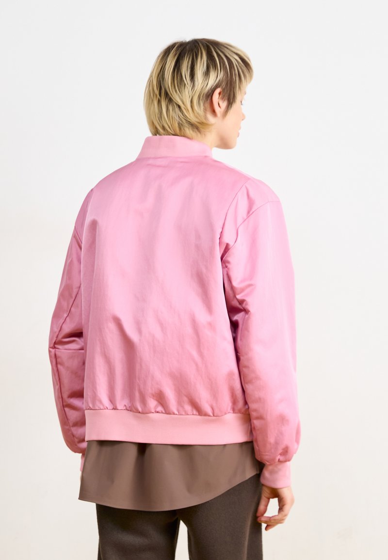Rosa Bomberjacke mit glatter Textur, gerippten Bündchen und Saum sowie einem minimalistischen Design, von der Rückseite mit einem schichtartigen braunen Oberteil darunter gezeigt.