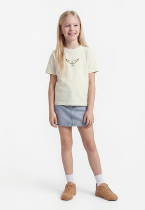 Fille souriante aux longs cheveux blonds portant un t-shirt crème avec un motif d'ailes, une jupe en denim clair, des chaussettes blanches et des baskets marron, debout sur un fond blanc.