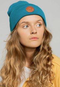 Turkusowa, prążkowana czapka beanie z brązową okrągłą naszywką z motywem góry. Modelka ma długo falujące, blond włosy i nosi żółtą kurtkę.