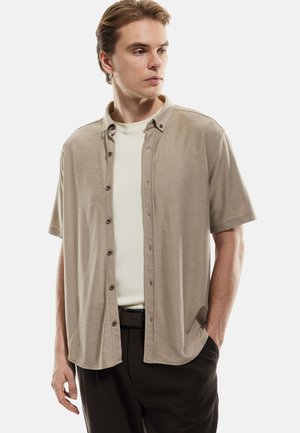Camisa de manga corta beige con cuello y rayas horizontales, abotonada al frente, llevada sobre una camiseta de color claro. Se ven pantalones oscuros y un cinturón.