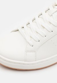 Baskets en cuir blanc avec surface texturée, lacets plats blancs et semelle en caoutchouc beige contrastante. Design minimaliste avec détails cousus.