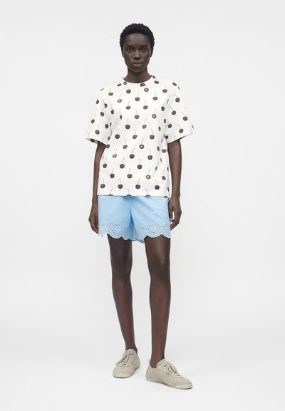 Γυναίκα στέκεται φορώντας λευκό μπλουζάκι με στάμπα κερασιών, ανοιχτό μπλε σορτς με κυματιστό τελείωμα και μπεζ αθλητικά παπούτσια σε απλό λευκό φόντο.