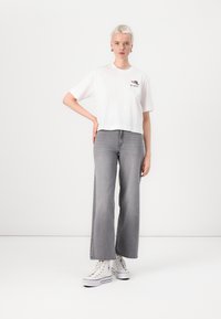 Hvit avkortet t-skjorte med grafisk trykk, kombinert med vide grå jeans og hvite joggesko. Enkelt design, myk stoff, avslappet passform.
