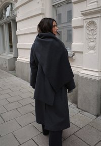 Tamsiai pilka vilnonė striukė su drabužiu ant nugaros, perdidelėmis rankovėmis ir švelnia tekstūra, papildyta kasdieniu įvaizdžiu ir miesto aplinka.