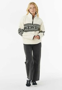 Fleece pullover in crème met een zwart geometrisch patroon over de borst, voorzien van een half-rits kraag en contrasterende zwarte mouwaccenten.