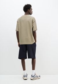 Oliven grøn tekstureret kortærmet skjorte parret med marineblå stribede shorts. Sneakers med sølv- og marineblå accenter, hvide sokker synlige.