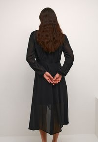 Černé mid-length šaty s dlouhými průhlednými rukávy a přiléhavým pasem, s volnou sukní, která částečně odhaluje nohy.