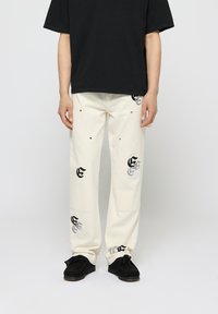Pantalones color crema con diseños gráficos en negro y plata, de corte estándar, con bolsillos y parte delantera plana. Combinados con una camisa negra.