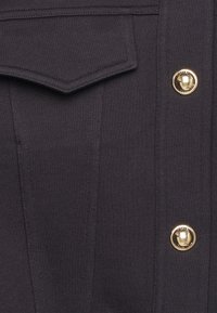 Tessuto navy scuro con una texture a coste verticali, dotato di una tasca sul petto e bottoni dorati. Design pulito senza ulteriori motivi.