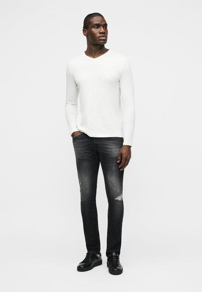 Άντρας που στέκεται, φοράει λευκό πουκάμισο με μακριά μανίκια, ξεθωριασμένο μαύρο τζιν με σκισίματα και μαύρα αθλητικά παπούτσια, μπροστά σε ένα απλό ανοιχτό φόντο.