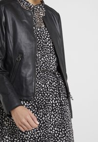 Černá kožená bunda s přiléhavým střihem, zipovým uzávěrem a bočními kapsami, navrstvená na šatě s černobílým puntíkovaným vzorem.
