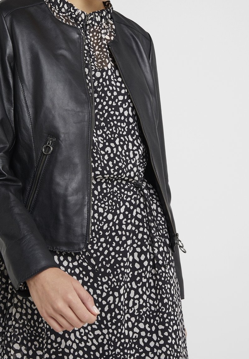 Černá kožená bunda s přiléhavým střihem, zipovým uzávěrem a bočními kapsami, navrstvená na šatě s černobílým puntíkovaným vzorem.