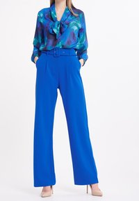 Blouse bleue avec un col à nœud et des motifs abstraits verts, associée à un pantalon bleu taille haute à jambes larges et des talons hauts nude.