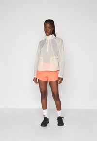 Veste légère et translucide avec un col haut, deux poches avant et des accents réfléchissants ; associée à un short orange vif et des baskets noires.