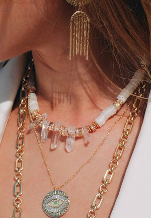 Drei Halsketten: eine mit klaren Kristallen und goldenen Akzenten, eine mit einem runden Anhänger mit einem Augendesign und eine dicke goldene Kette.