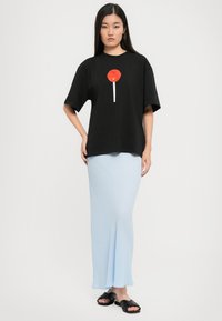 Kvinna i en svart oversized t-shirt med en röd klubba-graphic och en ljusblå lång kjol, stående mot en enkel vit bakgrund.