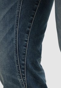 Close-up of dark blue denim jeans showing fabric texture and vertical seam detail along the leg against a plain background.