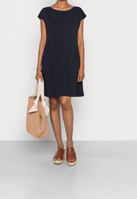 Laivastonsininen mekko, jossa on småärmelöitä ja löysä muoto, yhdistettynä ruskeisiin punottuihin platoforinsandaaleihin ja beigelle olkalaukulle, jossa on solmittavat kahvat.