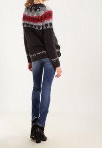 Suéter de punto negro con un cuello de diseño en rojo y gris, llevado sobre jeans azules ajustados y botas de tacón negro, visto desde la parte posterior.