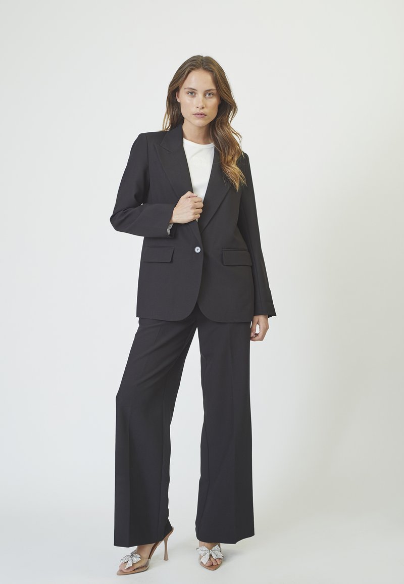 Black tailored suit with a fitted blazer featuring notch lapels and two front pockets, paired with wide-leg trousers and embellished heels.