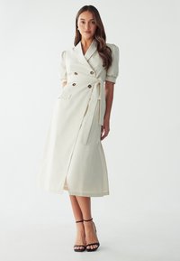 Vestido envolvente de color crema con mangas cortas abullonadas, parte delantera con doble botón, cinturón en la cintura y una abertura alta en el muslo. Llevado con tacones negros de tiras.