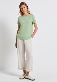 Vaaleanvihreä puuvillapaita, jossa on pyöreä pääntie ja yksinkertainen valkoisesta tekstistä koostuva graafinen kuvio. Yhdistetty vaaleisiin leveälahkeisiin pellavahousuihin ja valkoisiin lenkkareihin.