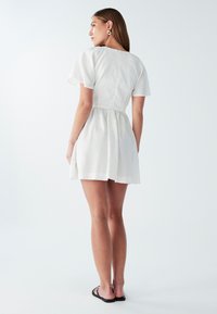 Biely krátky šaty s krátkymi puff rukávmi, zúženým pásom a rozšírenou sukňou. Tkanina vyzerá ľahko a má štruktúru, vhodná na teplé počasie.