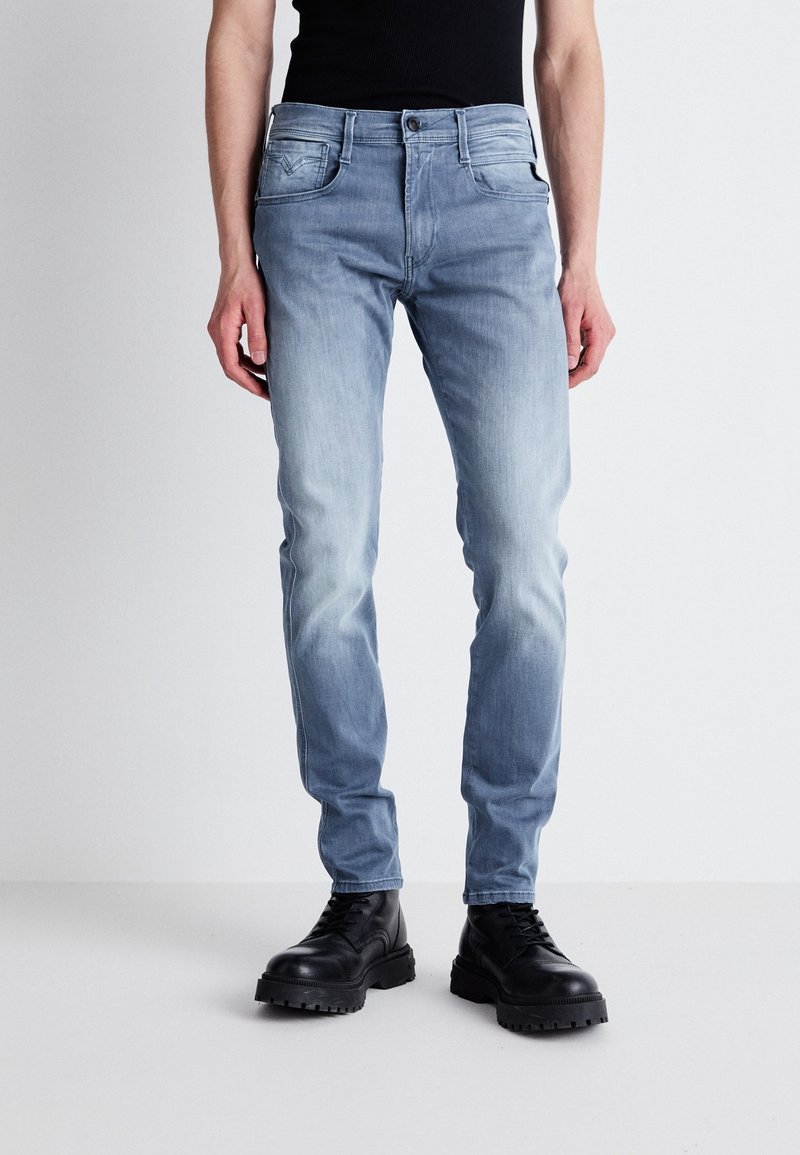 Personne portant un jean slim bleu clair et des bottes noires épaisses, debout devant un fond blanc uni.
