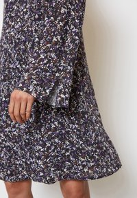 Blumenkleid mit langen, weit ausgestellten Ärmeln. Schwarzer Stoff mit kleinen weißen, lila und braunen Blumen. Leichte, fließende Textur. Der Saum endet über dem Knie.