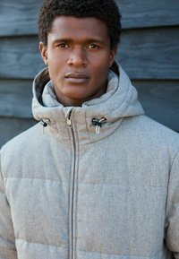 Grau gequiltete Jacke mit hohem Kragen und verstellbaren Kordelzügen. Verfügt über einen Frontreißverschluss und einen glatten, strukturierten Stoff.