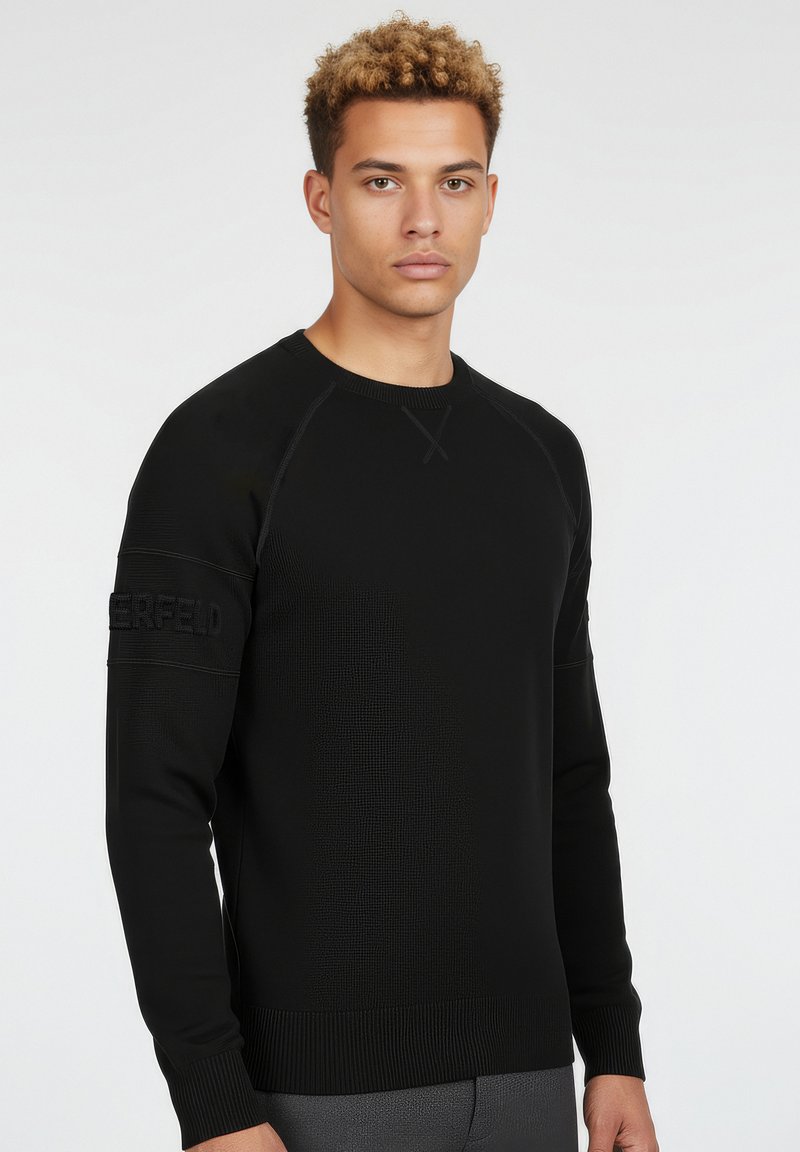 Nuori mies, jolla on kiharat hiukset ja joka pukeutuu mustaan pitkähihainen neuleeseen, jossa on kohopainettu teksti hihoissa, seisoo yksinkertaisen taustan edessä.