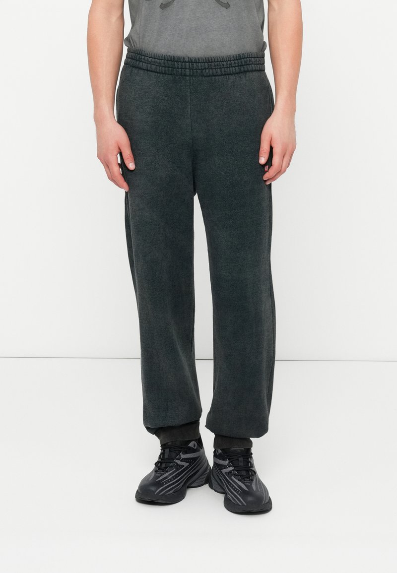 Person, der dunkelgraue Fleece-Jogginghosen und schwarze Sportschuhe trägt, steht vor einem schlichten weißen Hintergrund.
