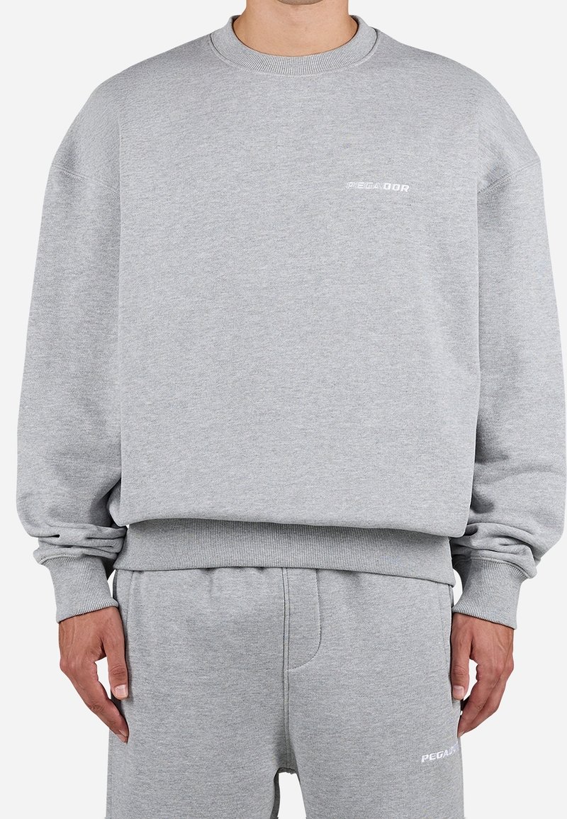 Homme portant un ensemble sweat-shirt et pantalon de survêtement assortis gris clair avec de petits logos blancs "PEGADOR" sur la poitrine et la cuisse.