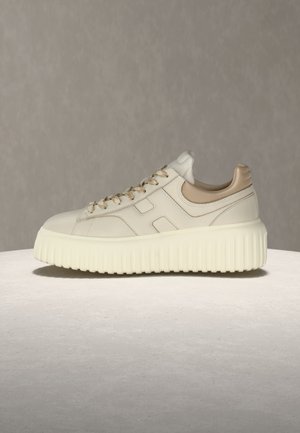 Sneaker in pelle color crema con suola spessa a coste, tallone imbottito beige e lacci abbinati su una superficie e uno sfondo neutri.