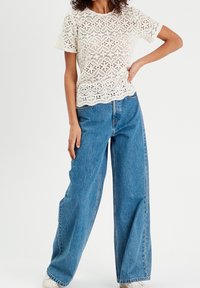 Haut crème en tricot de dentelle à manches courtes, associé à un jean en denim bleu moyen à coupe ample. Les deux articles présentent une silhouette décontractée et une texture douce.