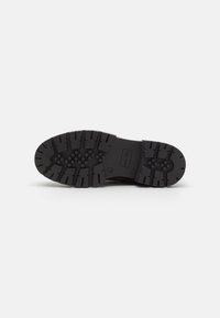 Semelle en caoutchouc noir avec des motifs de structure profonde et des surfaces texturées, présentant un logo de marque en relief et un design robuste pour une meilleure traction.