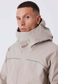 Chaqueta beige con capucha y cuello alto, con una textura suave y detalles grises en el pecho, diseñada para protección contra el clima.