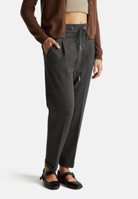 Tmavě šedé tepláky s elastickým pasem na šňůrku, bočními kapsami a volným střihem, doplněné černými botami se strapcovým designem.