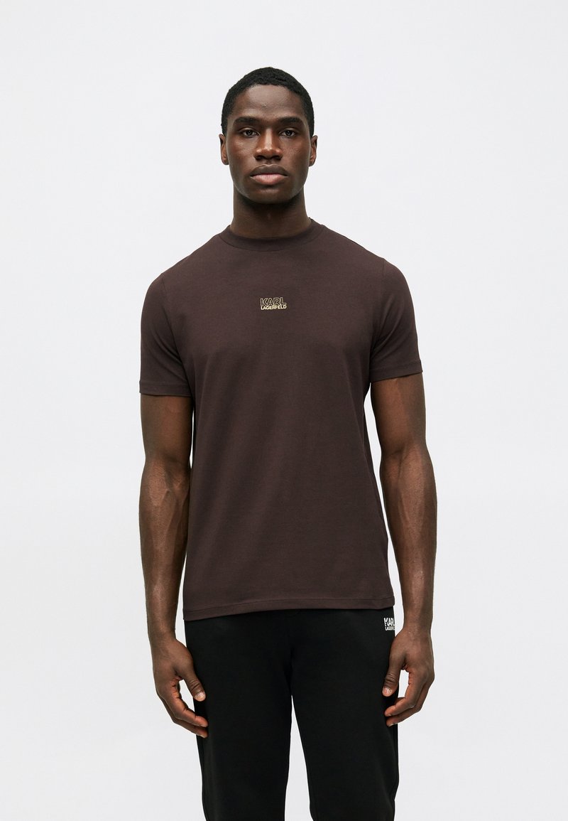 Tumši brūns kokvilnas T-krekls ar īsām piedurknēm, apaļu kakla izgriezumu un nelielu izšūtu logotipu uz krūtīm. Gluda tekstūra, pieguļošs dizains.