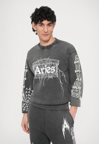 Temno siva jopa z belimi grafičnimi potiski, vključno z pajčevinami in napisom. Ima okrogel izrez in rebraste manšete, stajling z ujemajočimi spodnjimi deli.