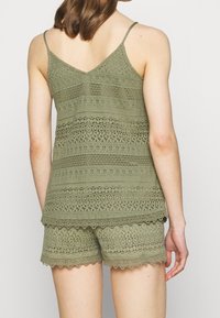 Olivově zelený dvoudílný outfit obsahuje krajkový top na ramínka a šortky s vlnitými okraji, které ukazují texturovaný, vzorovaný design látky.