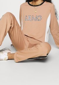Liu Jo souprava na volný čas světle béžové barvy z bavlny. Tričko s dlouhým rukávem a černým výstřihem, ozdobené třpytivým logem; kalhoty se shodnými černými akcenty a pruhy.